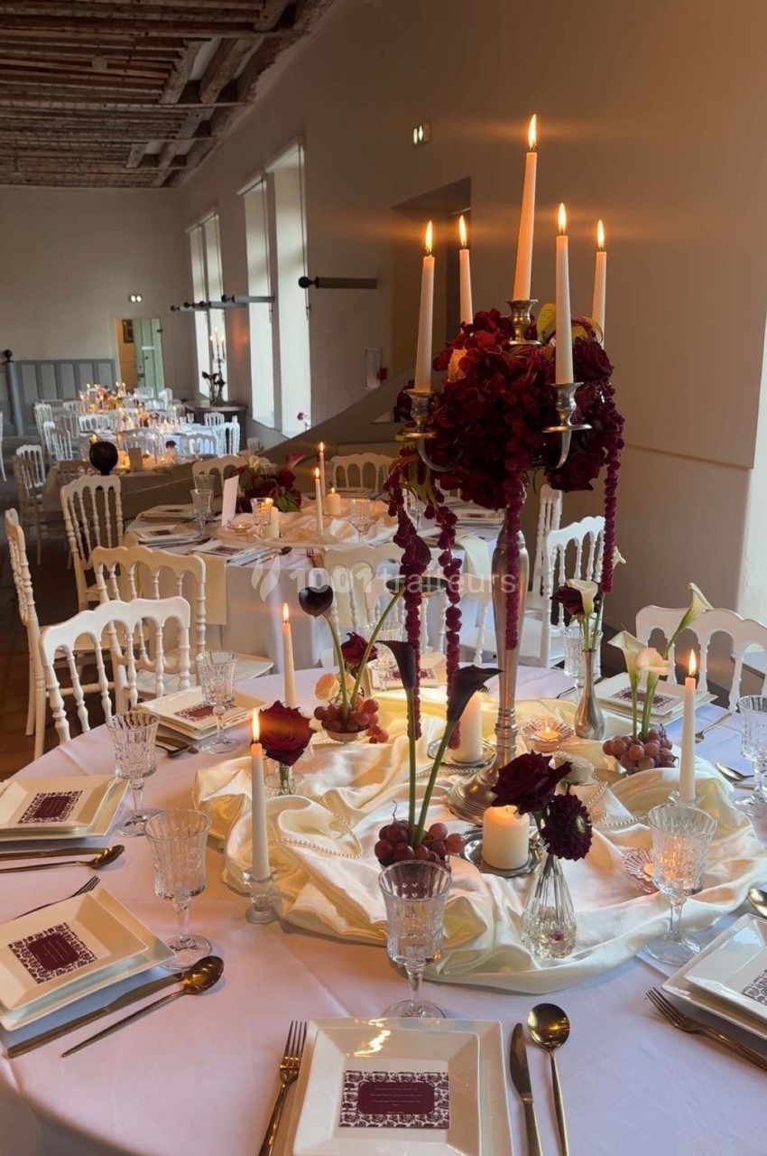 Table de réception élégamment dressée avec chandeliers, fleurs pourpres et vaisselle raffinée dans une salle lumineuse.