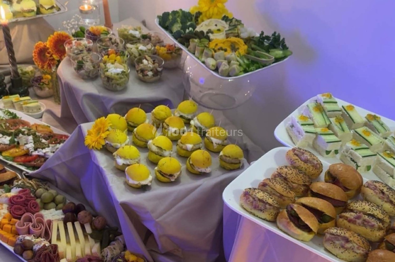 Buffet avec divers plats, dont sandwichs, salades, fruits et plateaux de fromage, décoré de fleurs jaunes.