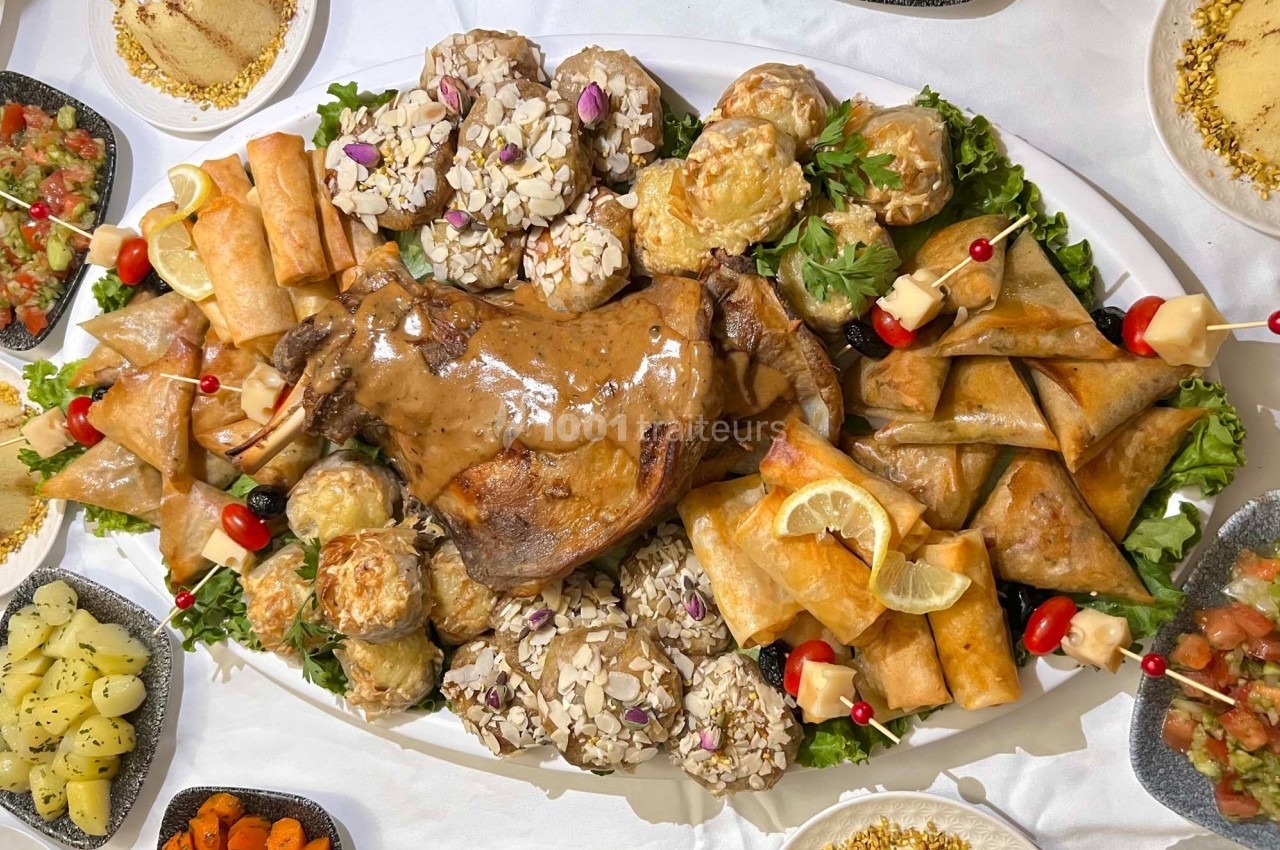 Plateau de méchoui entouré de briouates, feuilletés, rouleaux et garnitures variées sur une table dressée.
