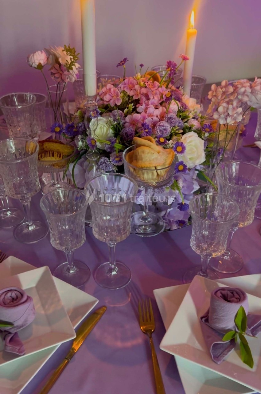 Table dressée avec vaisselle en verre, serviettes pliées, bougies allumées et centre de table floral dans des tons pastel.