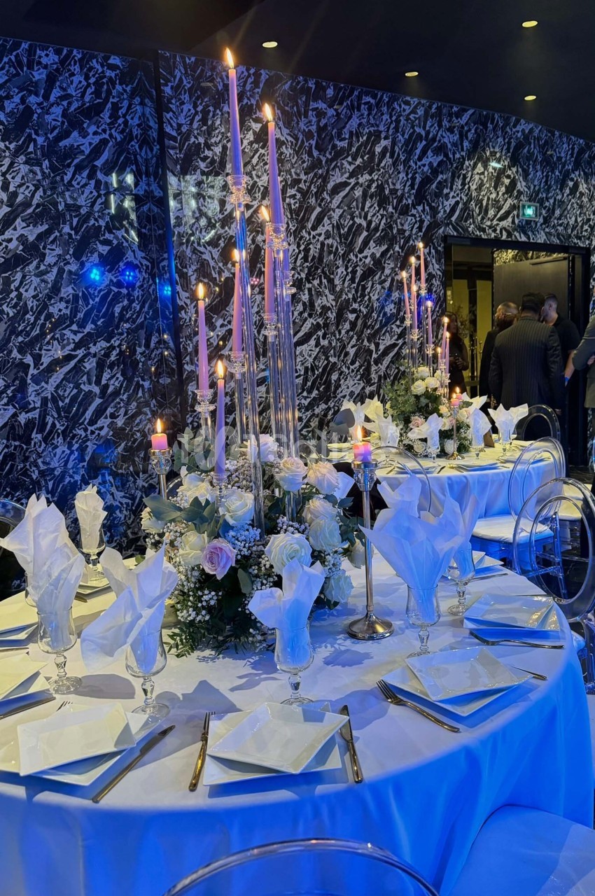Table élégamment dressée avec chandeliers, fleurs blanches et serviettes pliées, dans une salle au décor sombre et marbré.