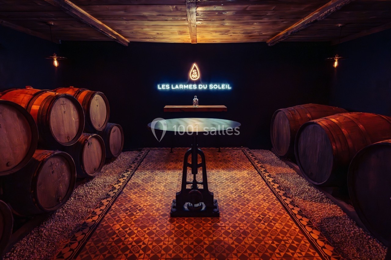 Salle de dégustation avec tonneaux en bois, table centrale et enseigne lumineuse ’Les Larmes du Soleil’ sur un mur sombre.