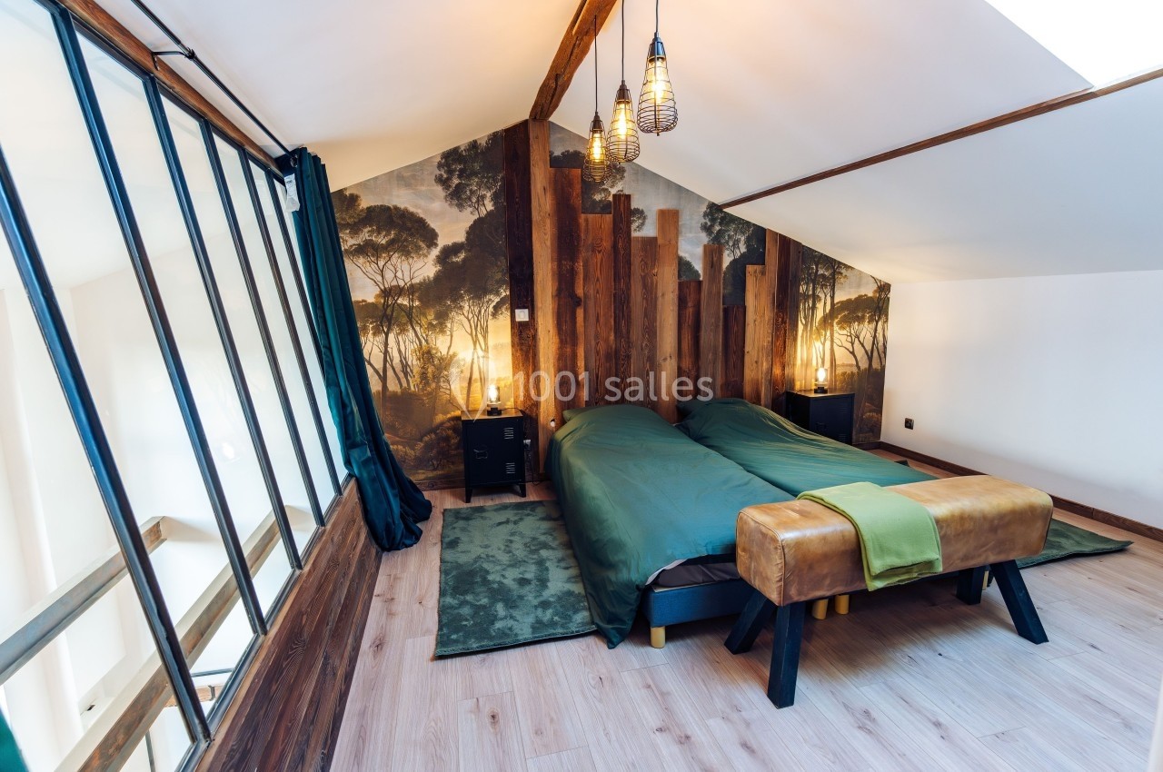 Chambre mansardée avec deux lits simples, décoration murale nature, poutres apparentes et éclairage suspendu.