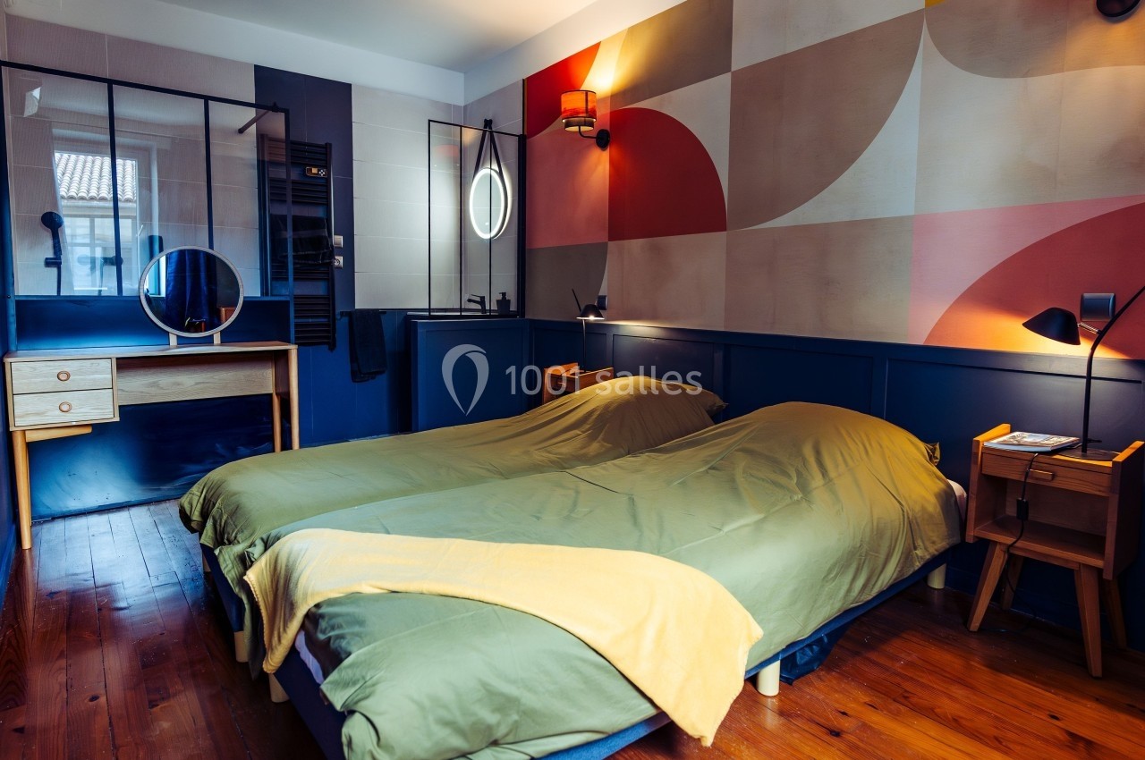 Chambre avec deux lits simples, murs colorés, bureau avec miroir, lampes de chevet et parquet en bois.