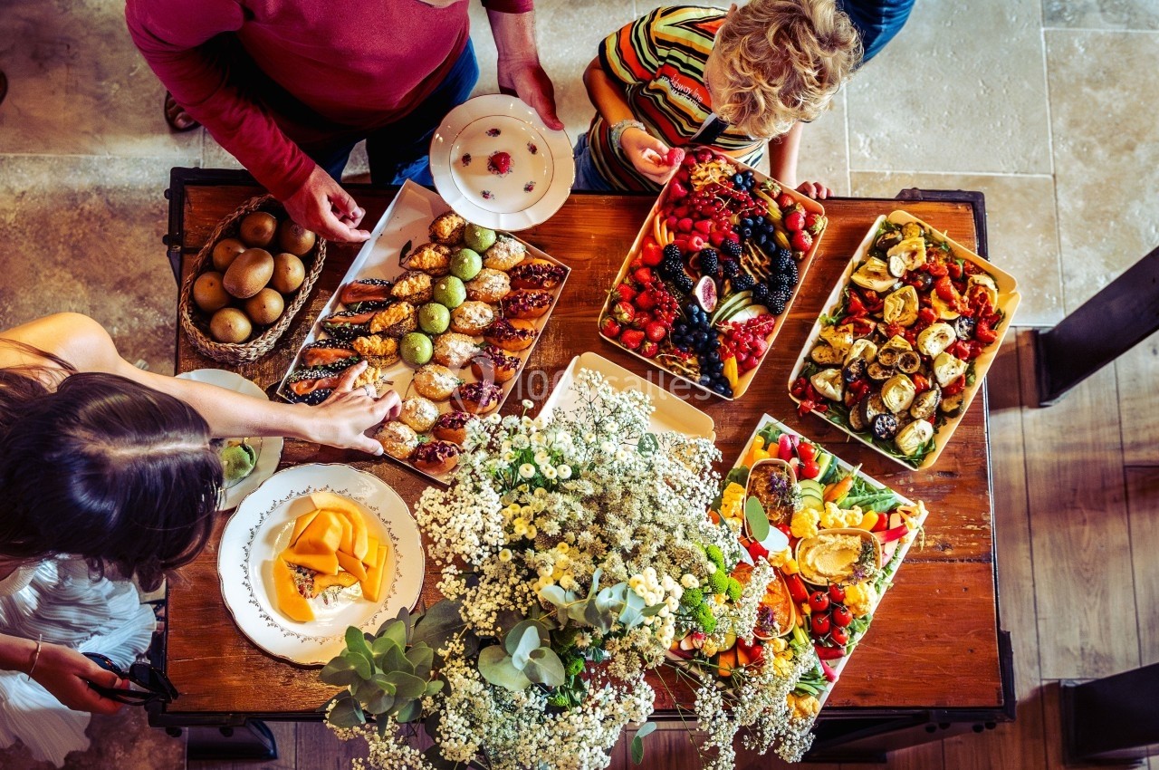 Vue plongeante sur une table garnie de plats variés, fruits, pâtisseries et fleurs, entourée de plusieurs personnes.