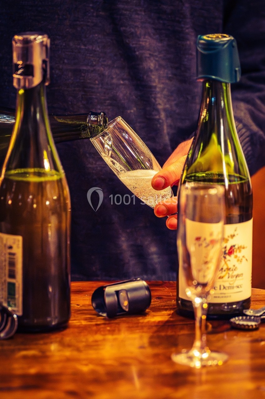 Une personne verse du vin pétillant dans une flûte sur une table en bois, entourée de bouteilles ouvertes.