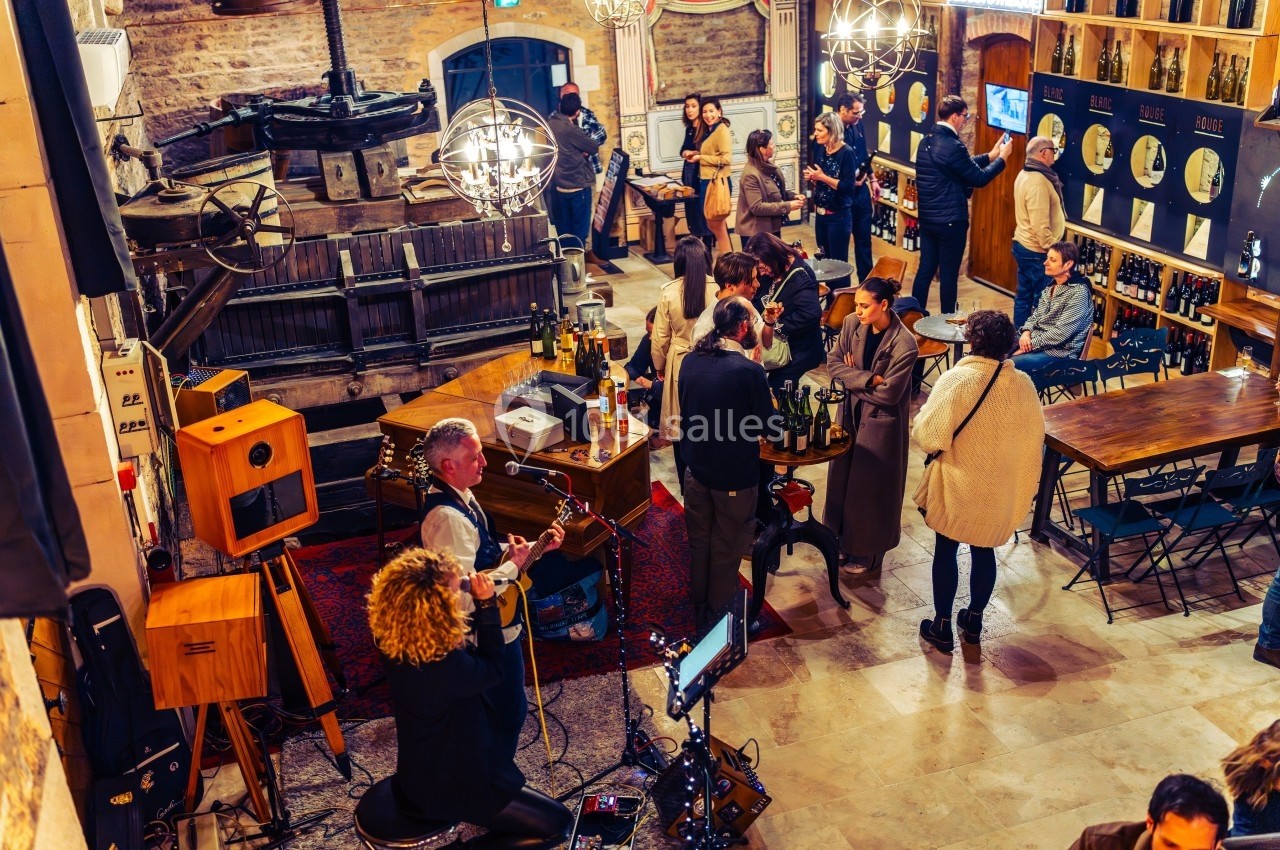 Groupe de musiciens jouant dans une salle animée avec des invités discutant autour de tables et d'un bar à vin.