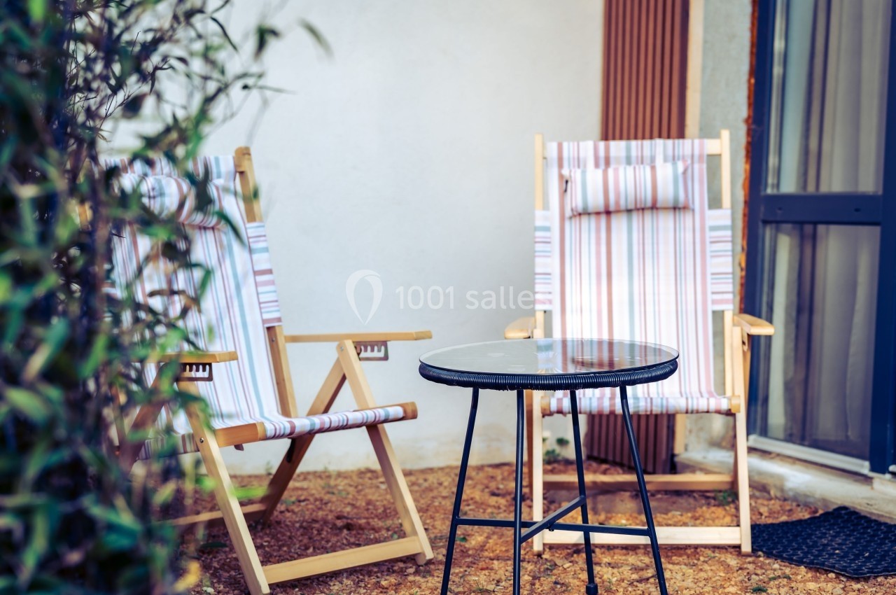 Deux chaises longues à rayures et une table ronde en verre sur une terrasse extérieure près d'une porte vitrée.