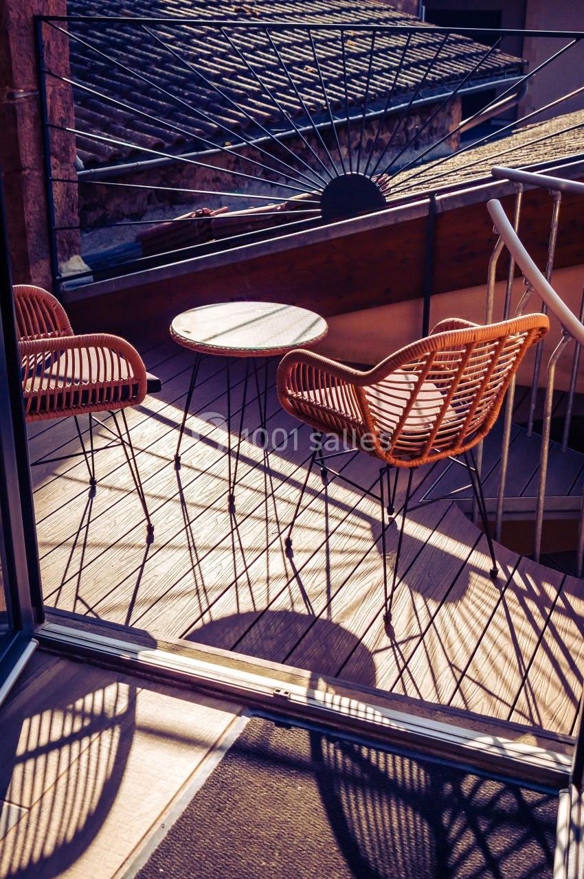 Deux chaises en rotin et une table ronde sur une terrasse ensoleillée avec des ombres marquées au sol.