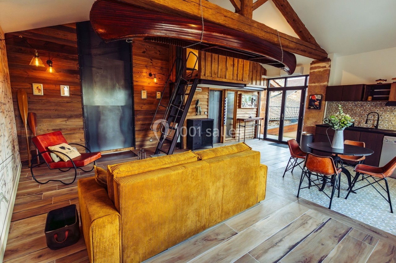 Salon chaleureux avec canapé jaune, mezzanine en bois, décoration rustique et cuisine ouverte dans un espace lumineux.