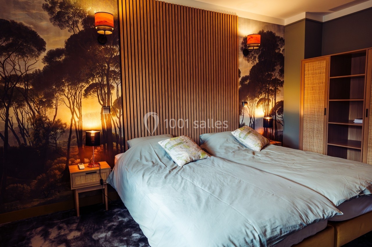 Chambre avec deux lits simples, tête de lit en bois, éclairage chaleureux et mur décoré d'une fresque forestière.