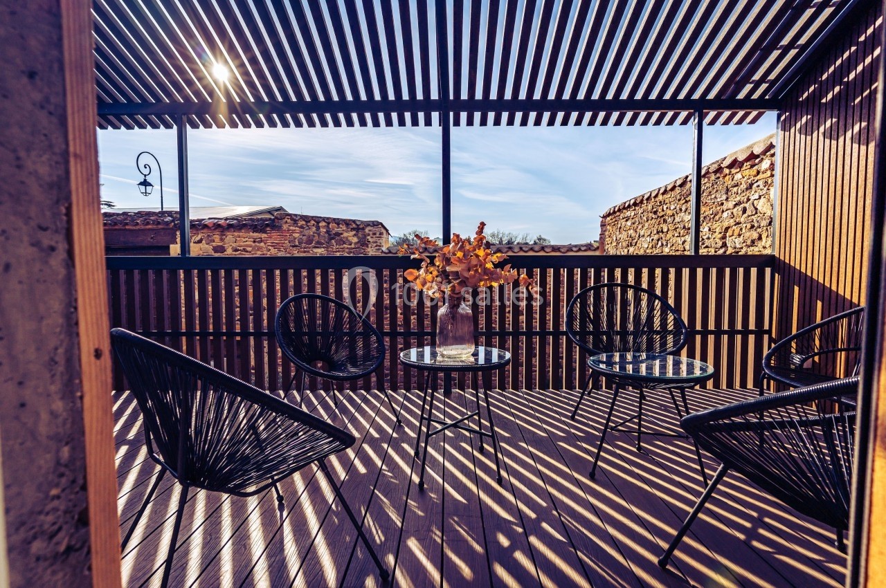 Terrasse en bois avec des fauteuils noirs, une table basse et un vase de fleurs, ombragée par une pergola à lames.