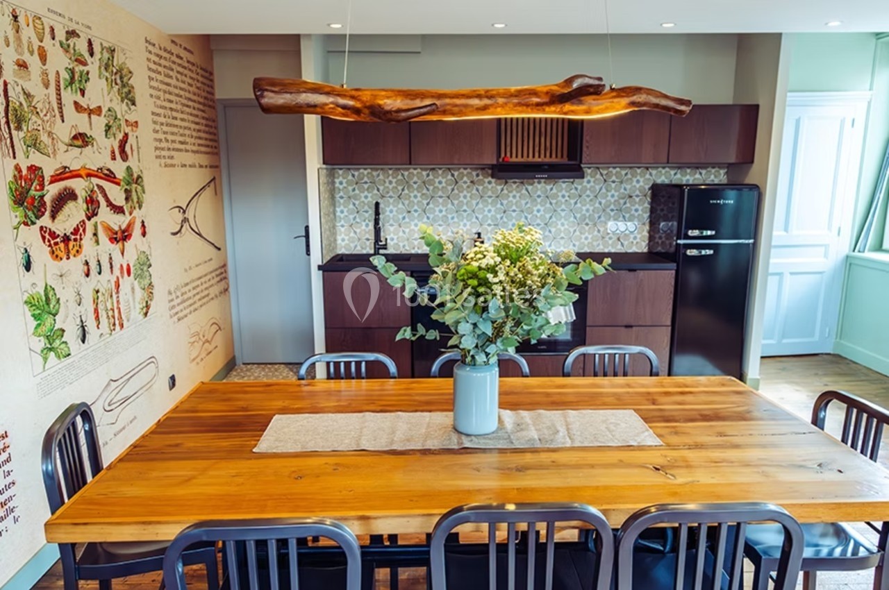 Salle à manger avec grande table en bois, chaises noires, cuisine ouverte moderne et décoration murale illustrée.