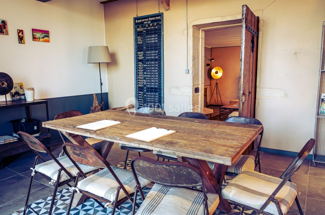 Salle de réunion avec table en bois, chaises métalliques, tableau mural et porte ouverte vers une pièce éclairée.