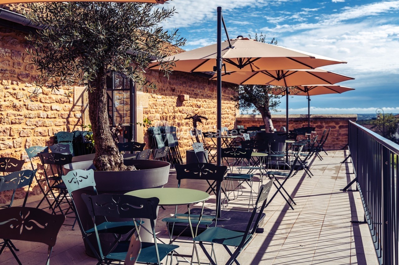 Terrasse ensoleillée avec tables, chaises en métal et parasols, bordée d'un mur en pierre et d'une rambarde.