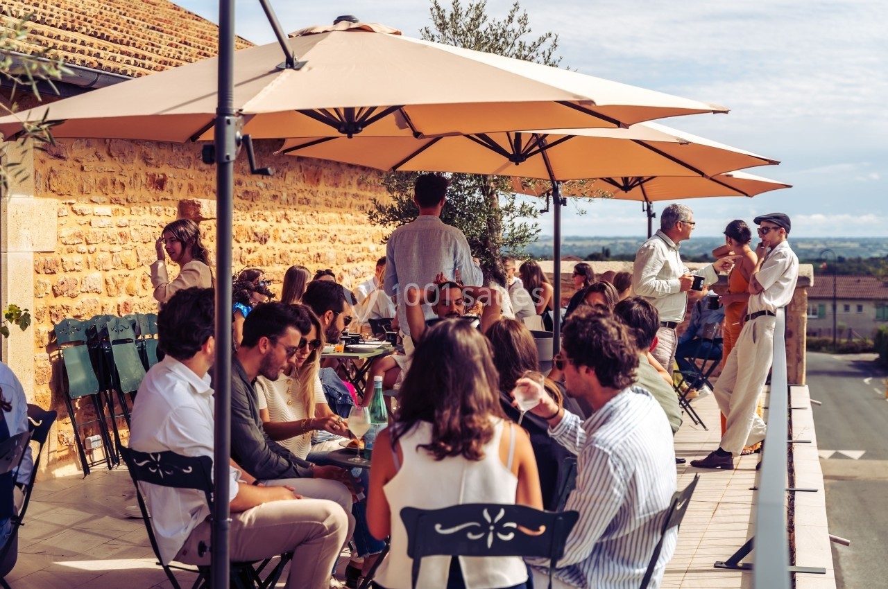Groupe de personnes assises et debout sur une terrasse ensoleillée, discutant sous de grands parasols beiges.