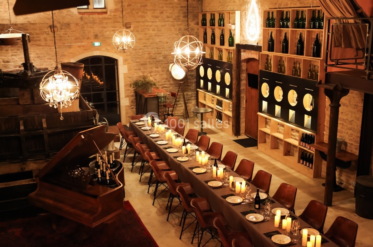 Salle chaleureuse avec une longue table dressée, des bougies allumées, un piano et des étagères de bouteilles de vin.