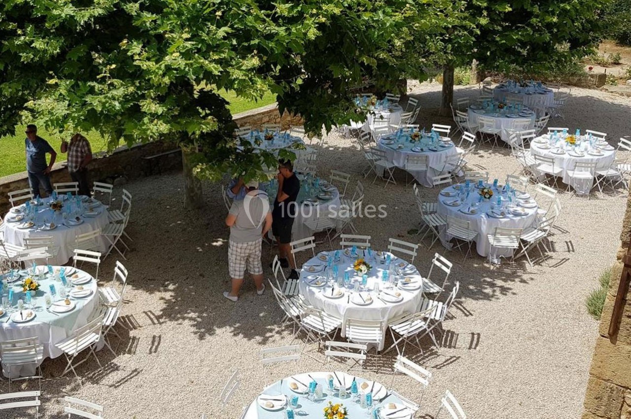 Tables rondes dressées avec nappes blanches et décorations florales, disposées en extérieur sous des arbres.