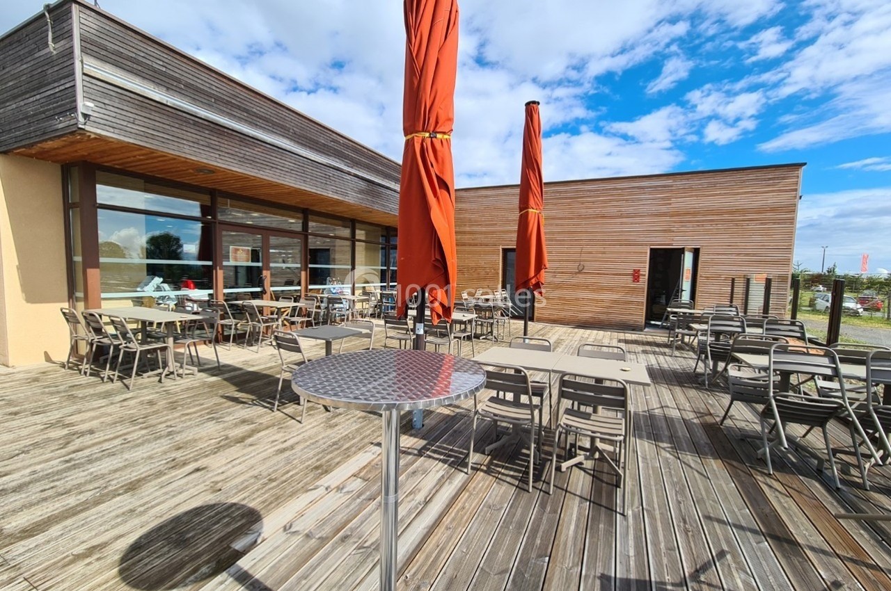 Location salle Gannat (Allier) - Paléopolis #5 Terrasse en bois avec tables, chaises et parasols rouges, devant un bâtiment moderne sous un ciel partiellement nuageux.