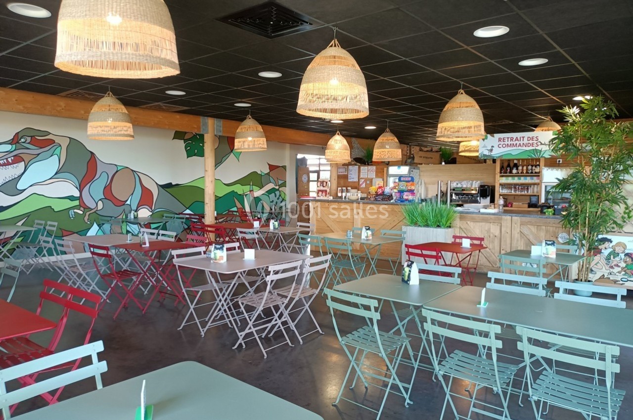 Location salle Gannat (Allier) - Paléopolis #7 Salle de restaurant lumineuse avec tables et chaises colorées, décor mural végétal et comptoir de commande au fond.
