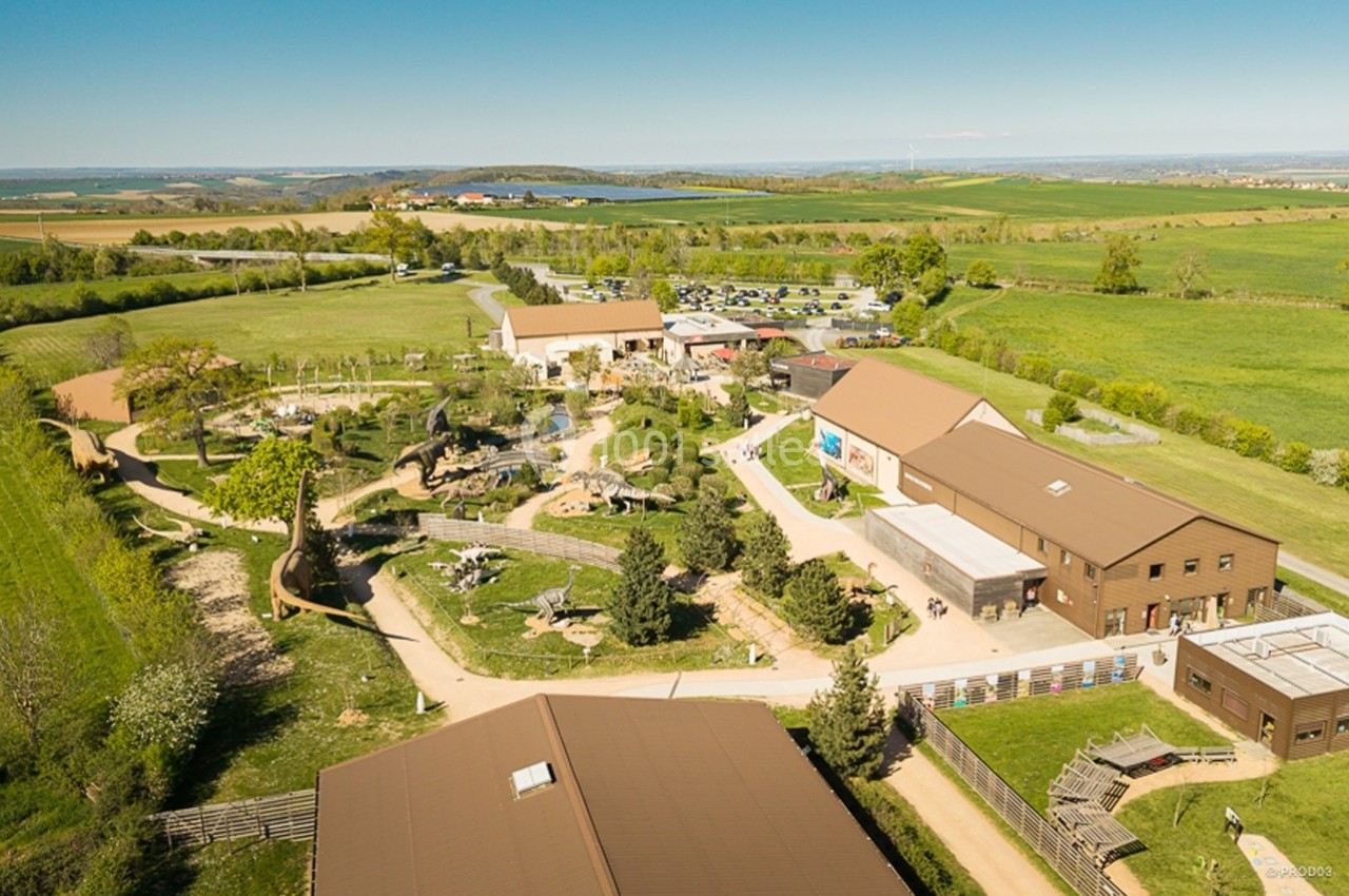 Location salle Gannat (Allier) - Paléopolis #1 Vue aérienne d'un parc avec bâtiments, espaces verts, chemins et parking en arrière-plan, entouré de campagne.