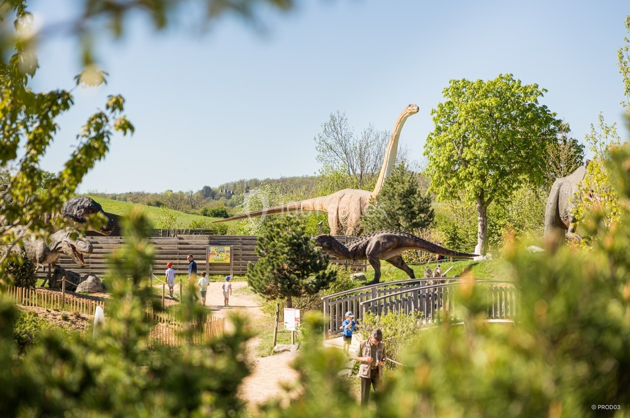 Location salle Gannat (Allier) - Paléopolis #16 Des statues de dinosaures grandeur nature dans un parc verdoyant, avec des visiteurs en promenade.