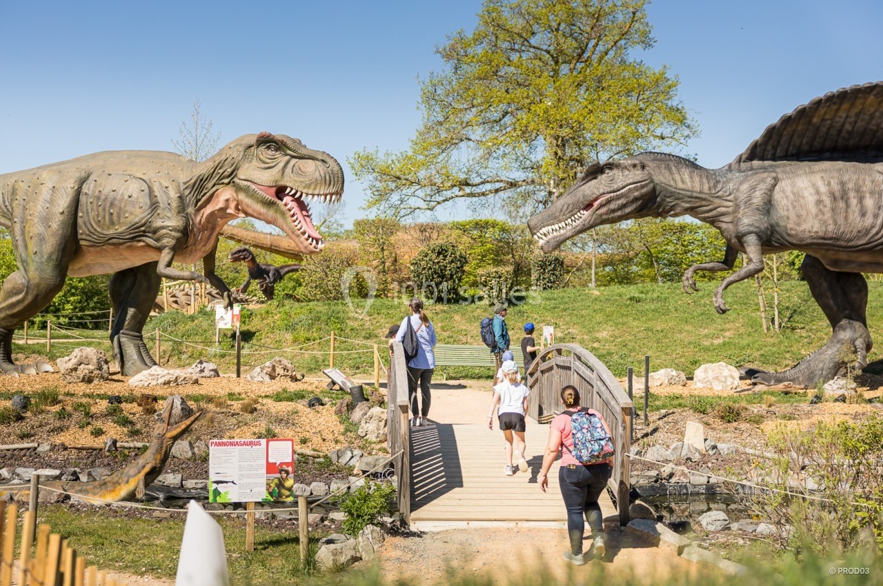 Location salle Gannat (Allier) - Paléopolis #18 Des visiteurs traversent un pont dans un parc avec des statues grandeur nature de dinosaures sous un ciel dégagé.