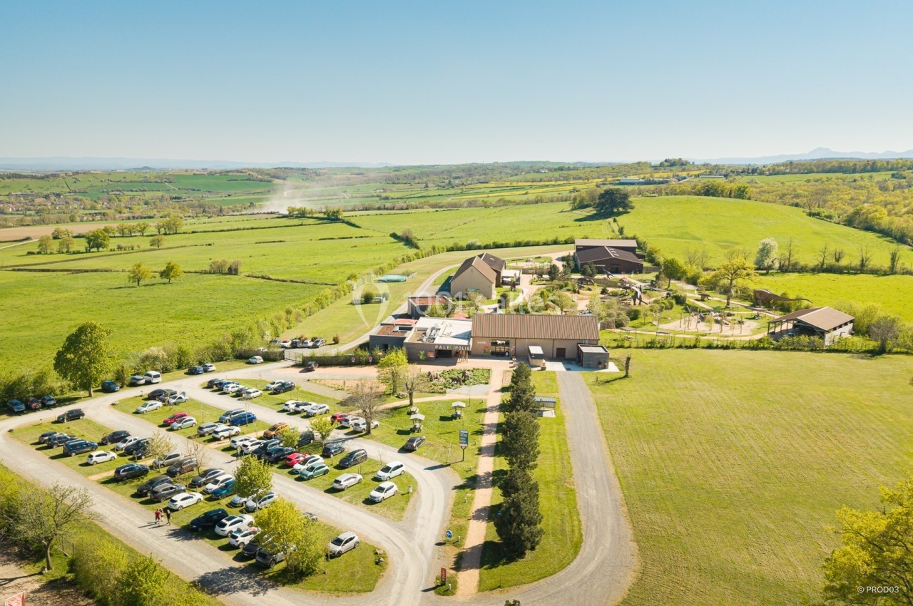 Location salle Gannat (Allier) - Paléopolis #10 Vue aérienne d'un complexe rural avec bâtiments, parking rempli de voitures et paysage verdoyant environnant.