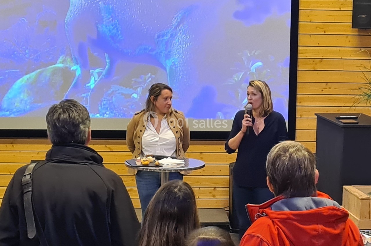 Location salle Gannat (Allier) - Paléopolis #3 Deux femmes parlent devant un public dans une salle avec un écran et des murs en bois.