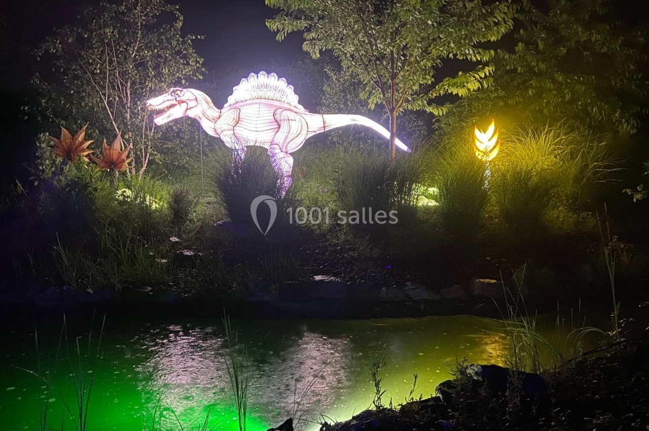 Location salle Gannat (Allier) - Paléopolis #20 Reproduction lumineuse d'un dinosaure près d'un étang éclairé, entouré de végétation et de lumières colorées.