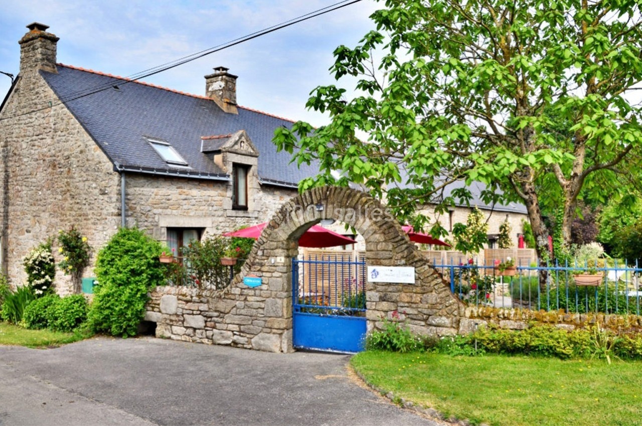 Façade d'une maison en pierre avec un portail en arche, un jardin arboré et des parasols colorés en arrière-plan.