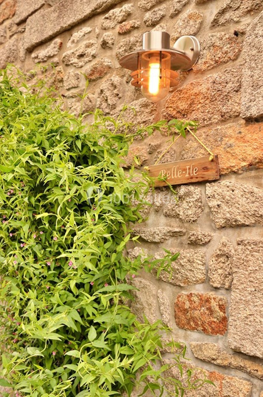Applique murale allumée sur un mur en pierre, entourée de verdure avec une petite pancarte en bois indiquant ’Belle-Île’.