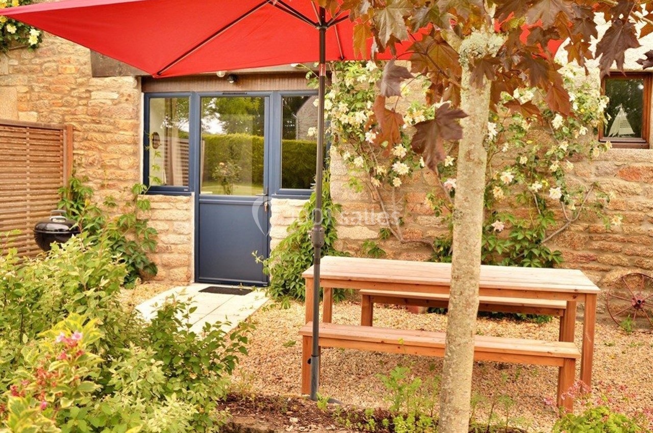 Entrée d'une maison en pierre avec porte bleue, table en bois sous un parasol rouge et jardin fleuri.