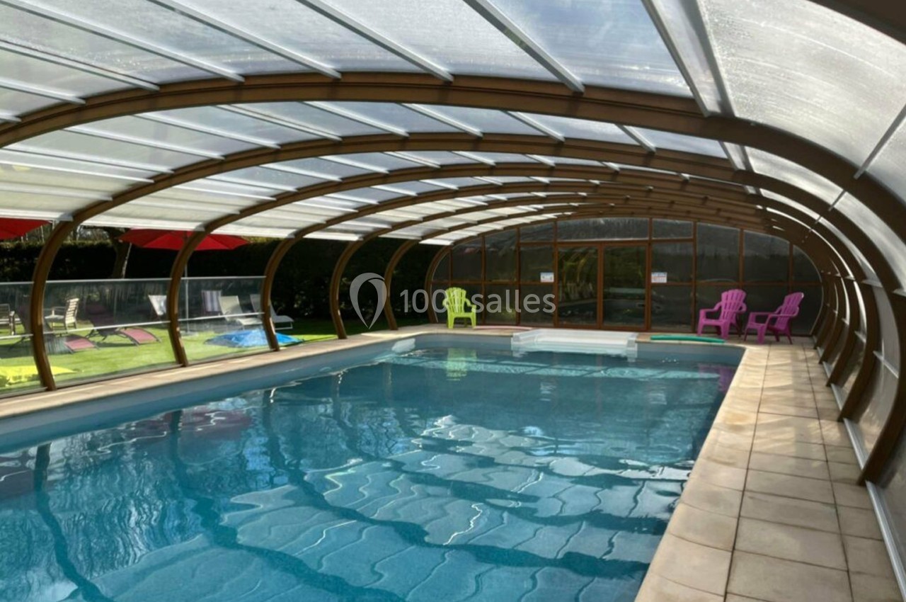 Piscine couverte avec structure en verre et métal, entourée de chaises colorées sur un sol carrelé et une pelouse.