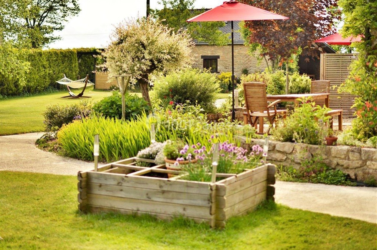 Jardin aménagé avec potager surélevé, table en bois sous parasols rouges et hamac en arrière-plan.