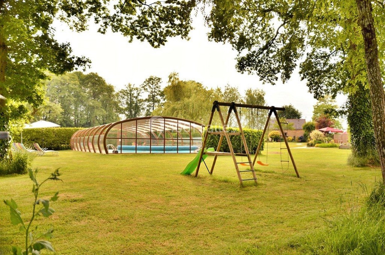 Jardin verdoyant avec balançoire en bois et toboggan, piscine couverte en arrière-plan entourée de végétation.