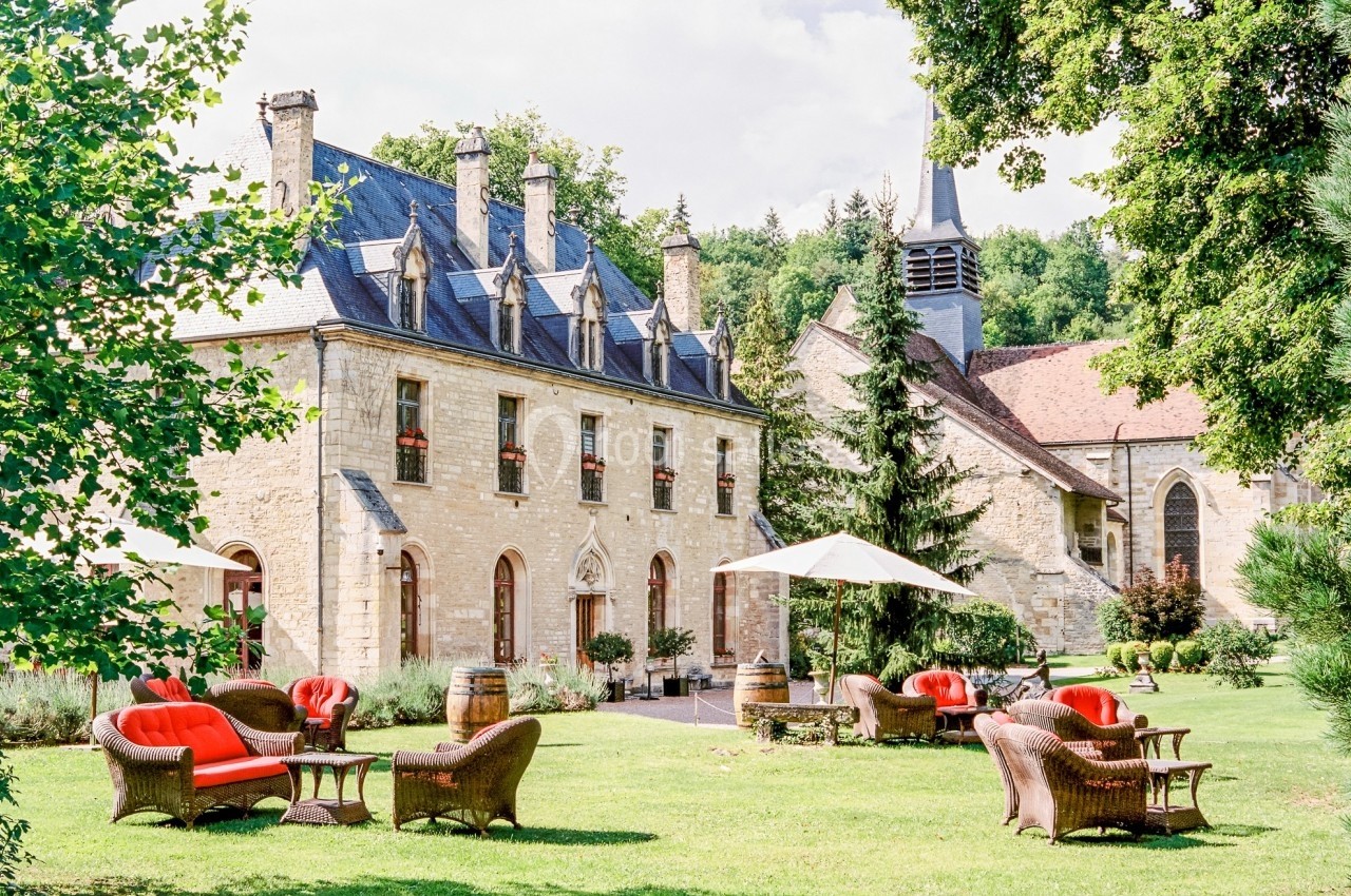 Jardin avec fauteuils en osier et parasols devant un bâtiment en pierre et une église dans un cadre verdoyant.