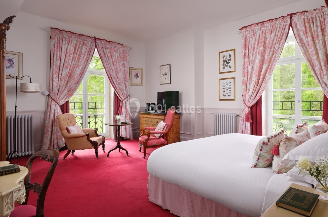 Chambre lumineuse avec lit double, fauteuils, bureau en bois, rideaux roses et fenêtres donnant sur un paysage verdoyant.
