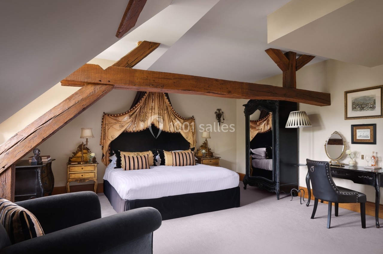 Chambre mansardée avec poutres apparentes, lit double, mobilier classique, bureau, miroir et fauteuils.