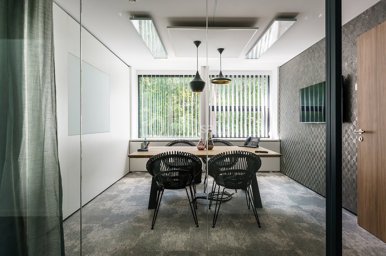 Salle de réunion moderne avec table, chaises noires, luminaires suspendus et grande fenêtre avec stores verticaux.