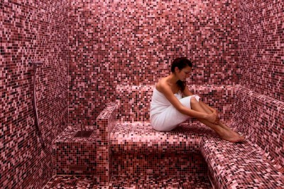 Miniature Le hammam de l'hôtel Dolce La Hulpe Brussels Femme assise dans un hammam aux carreaux roses, enveloppée dans une serviette blanche.