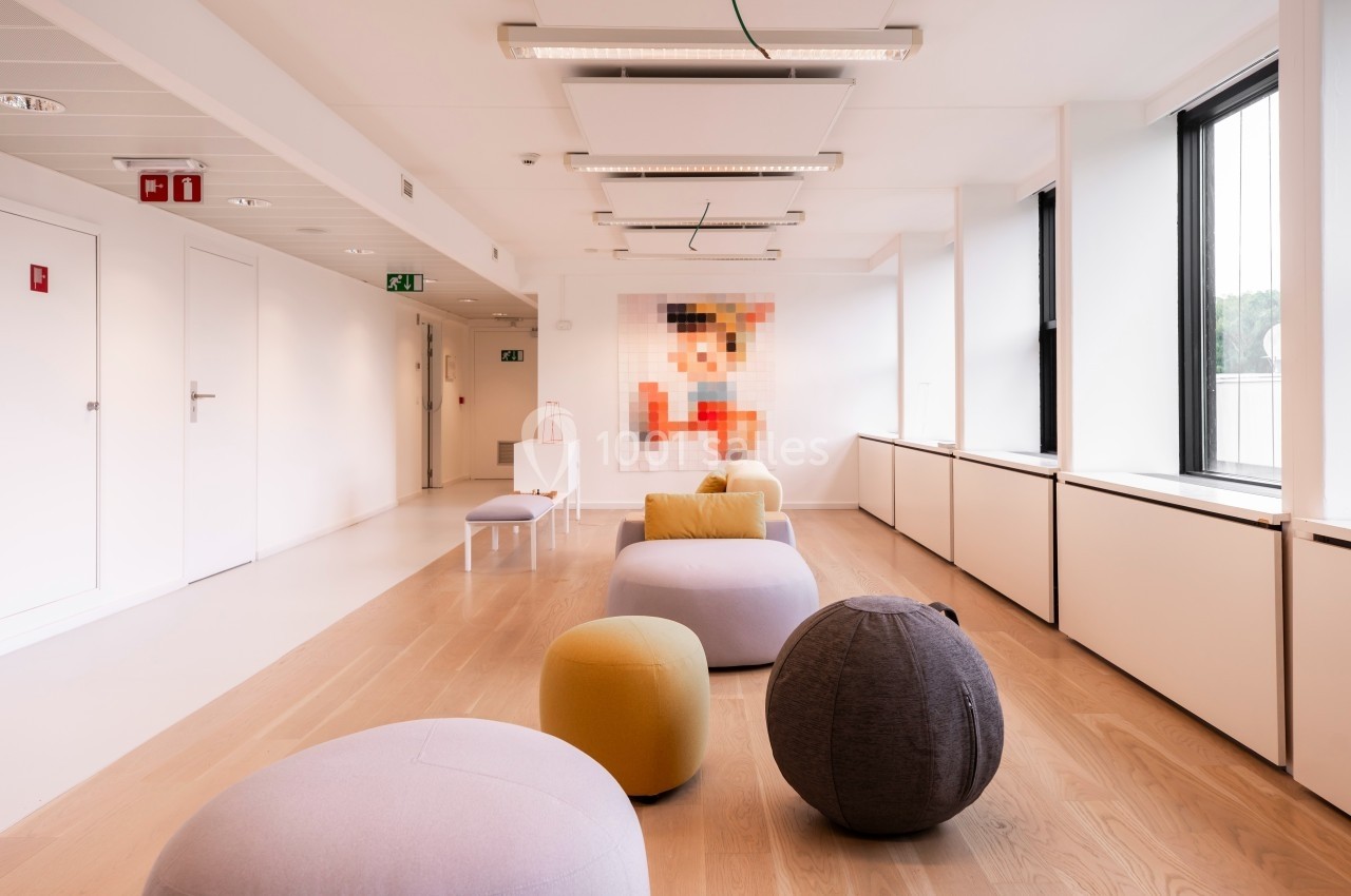 Espace lumineux avec des poufs colorés sur un sol en bois clair, murs blancs et une image floutée en arrière-plan.