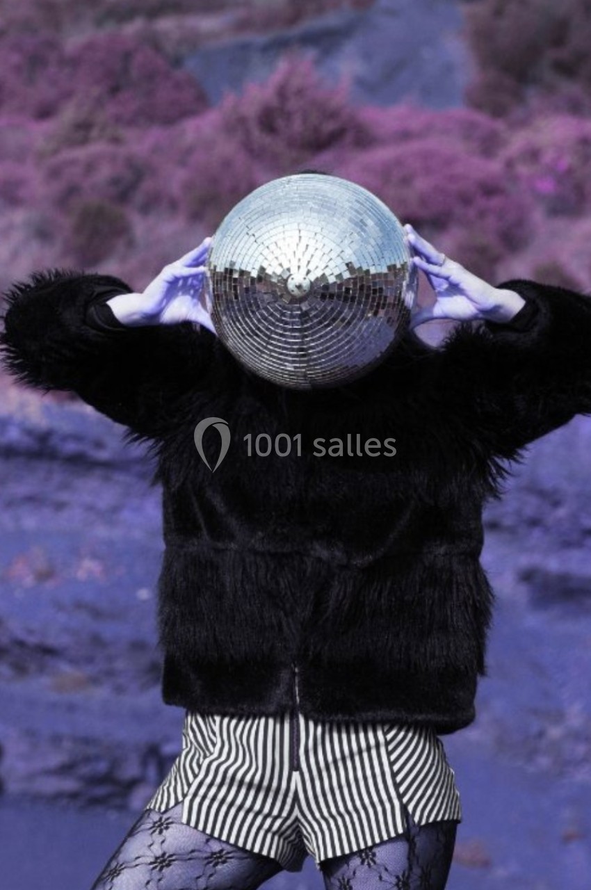 Personne portant une boule à facettes sur la tête, vêtue d'une veste noire et d'un short rayé, dans un paysage violet.