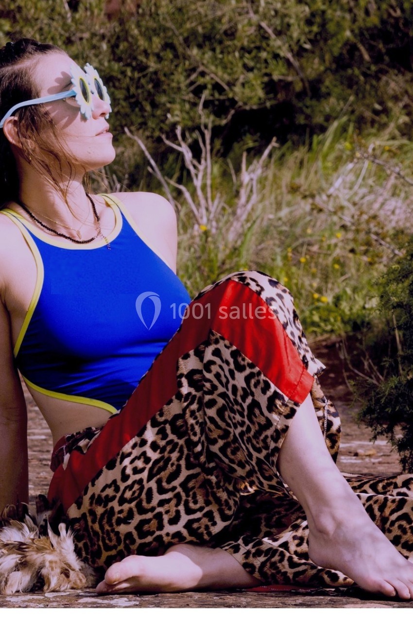 Femme assise en extérieur, portant un haut bleu, un pantalon à motif léopard et des lunettes fantaisie.