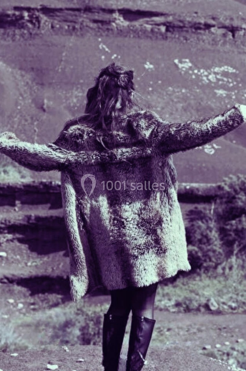 Femme de dos portant un manteau en fourrure, bras écartés, devant un paysage naturel avec des collines.