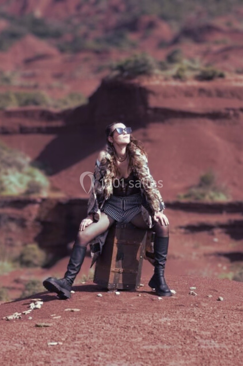 Femme assise sur une malle en plein air, entourée de collines rouges et de végétation clairsemée.