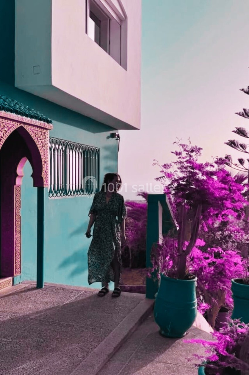 Une femme marche près d'une maison turquoise avec des plantes violettes et un ciel clair en arrière-plan.