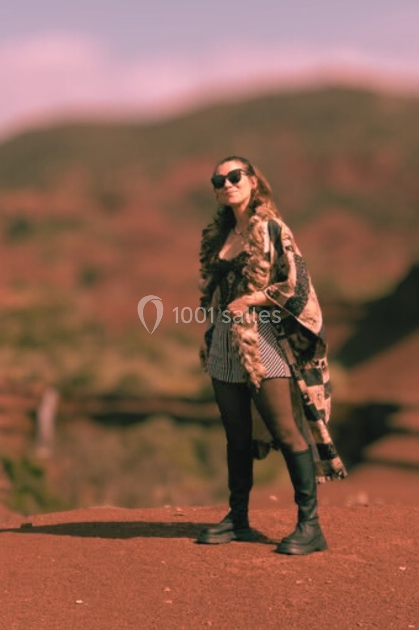 Femme debout sur un sol rouge, portant des lunettes de soleil et un manteau long, avec un paysage flou en arrière-plan.