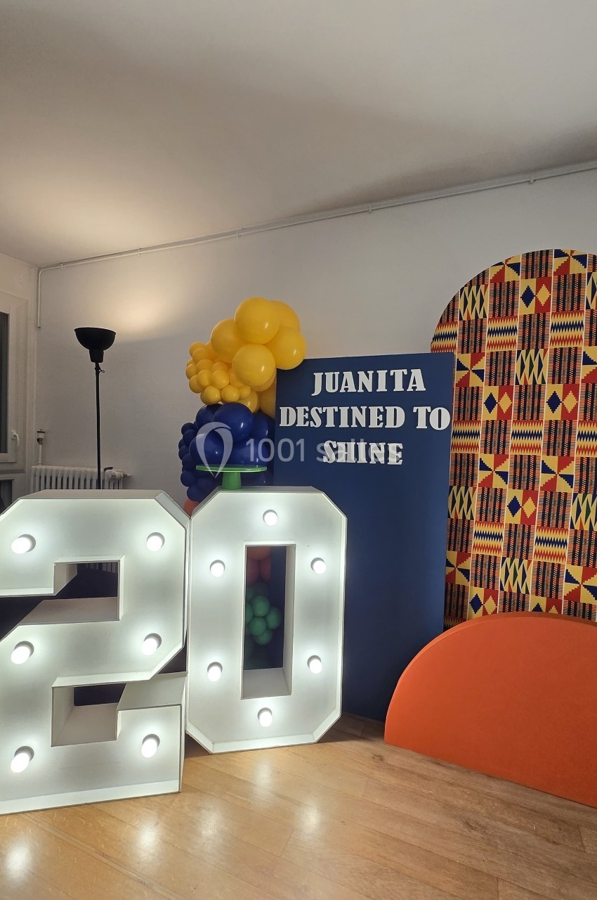 Décor festif avec un chiffre lumineux ’20’, des ballons colorés, un panneau ’Juanita Destined to Shine’ et un fond imprimé.