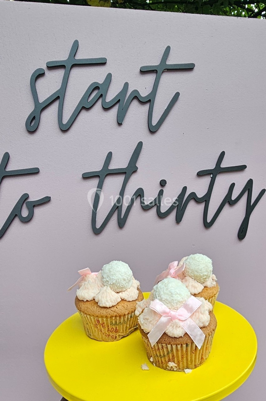 Des cupcakes décorés de crème et de rubans roses sont posés sur un présentoir jaune devant un panneau rose.